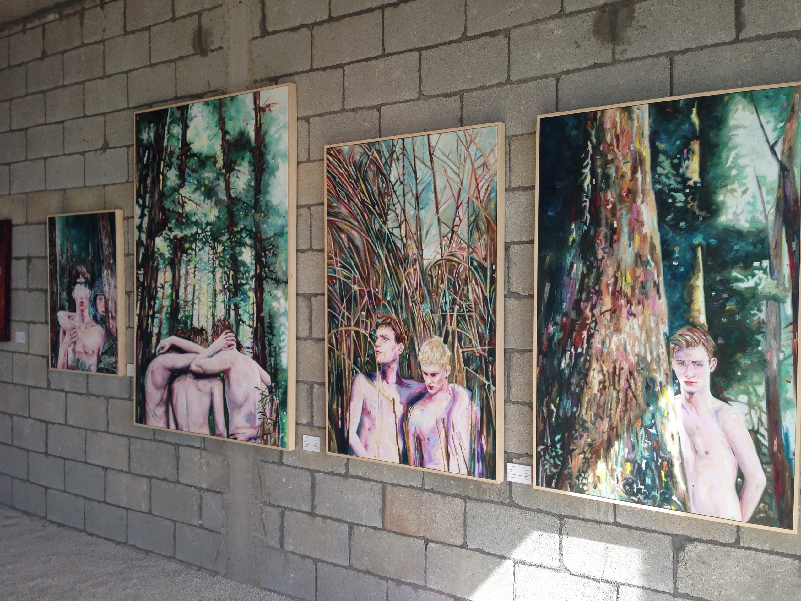 Angled installation view of five forest paintings displayed along an exposed block wall.