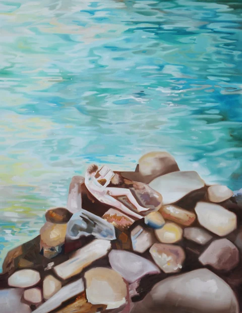 Figure partly hidden behind a pile of rocks beside turquoise water.
